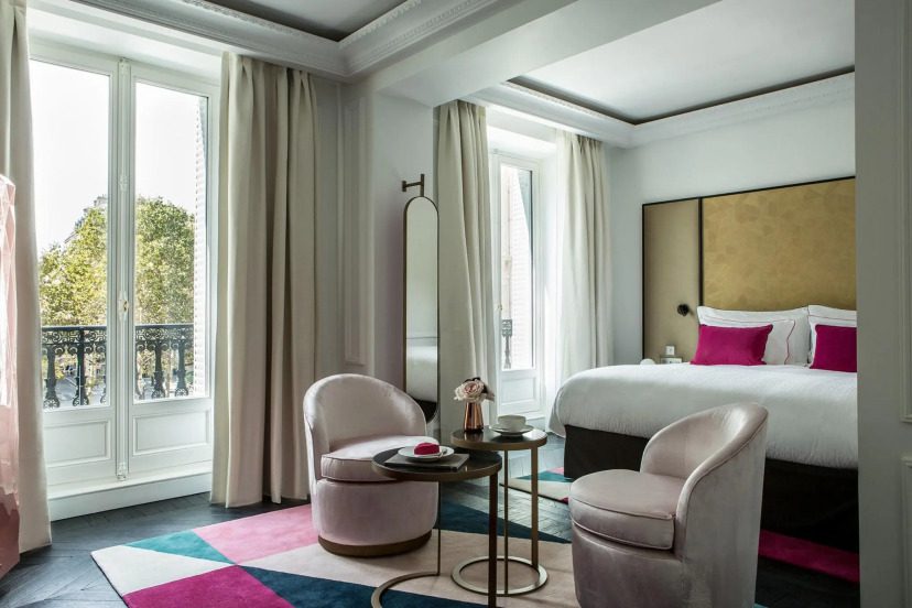 Elegant hotel room with large windows, soft beige curtains, and a luxurious bed with pink accents. Features plush chairs, a geometric rug, and a cozy ambiance.