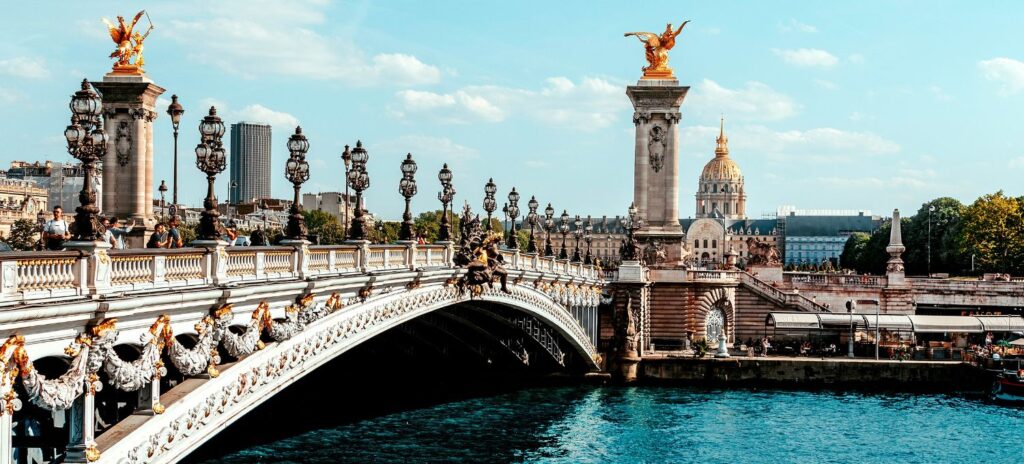 Elegant bridge with decorative lampposts and golden statues under a clear sky in Paris. A domed building is visible in the background.