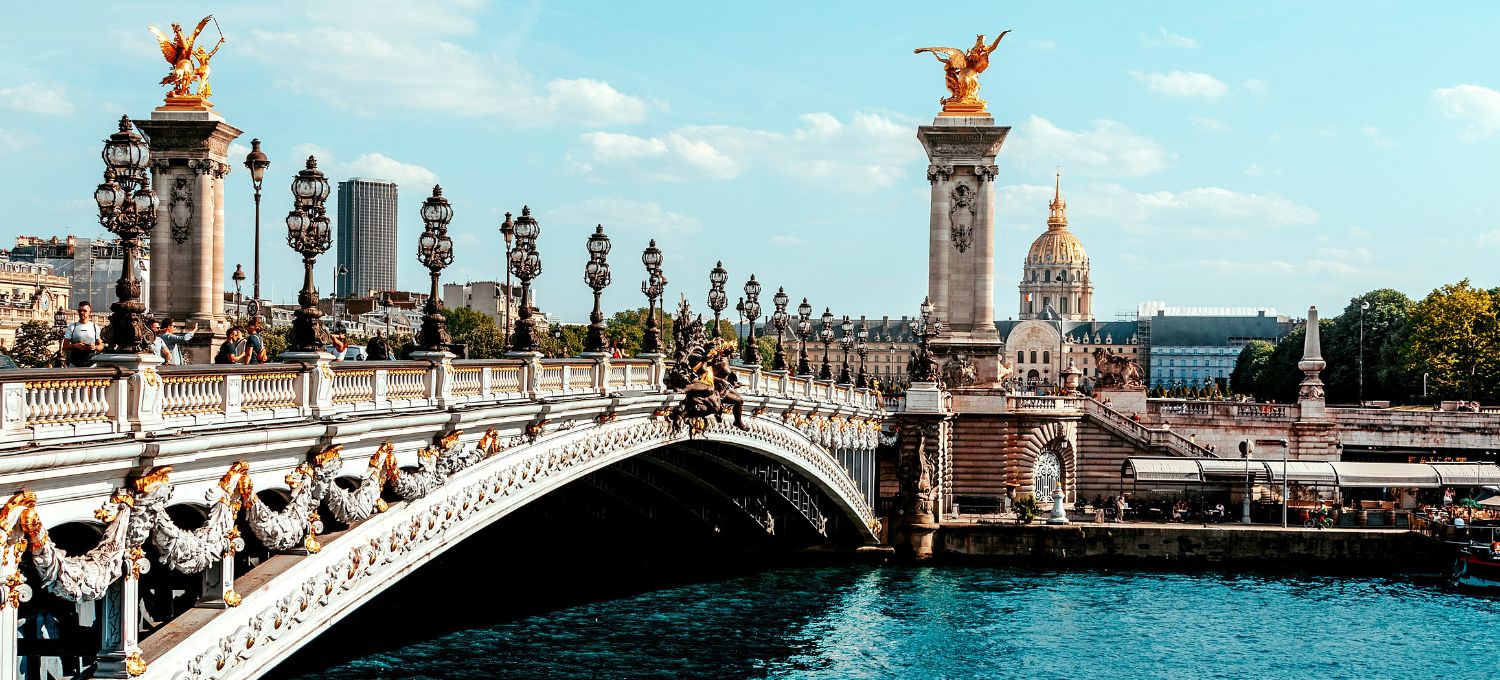 Elegant bridge with decorative lampposts and golden statues under a clear sky in Paris. A domed building is visible in the background.