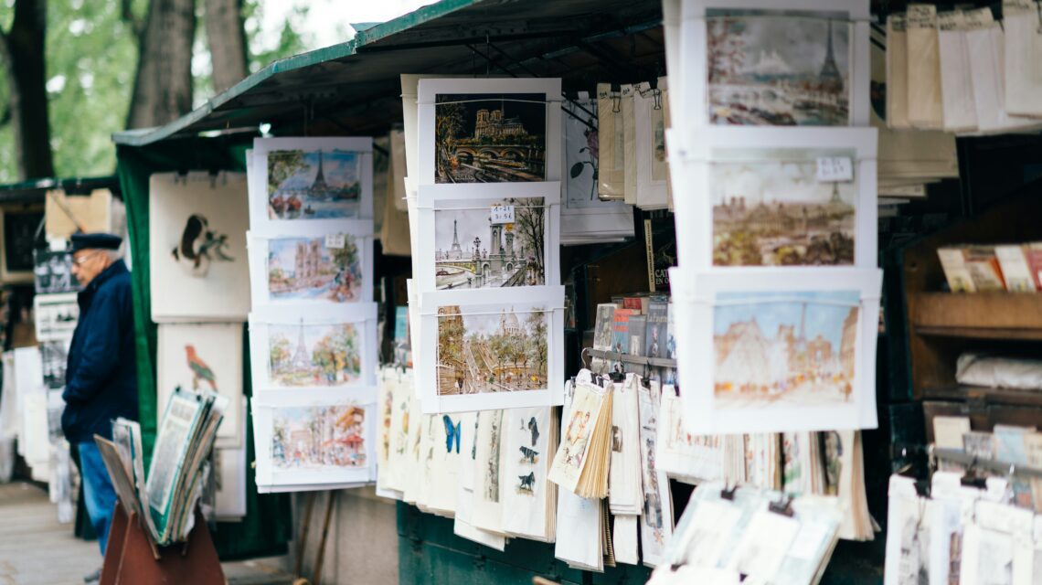 Outdoor art stall displaying colorful paintings and sketches of cityscapes and nature. An elderly person in a hat browses nearby. Relaxed atmosphere.