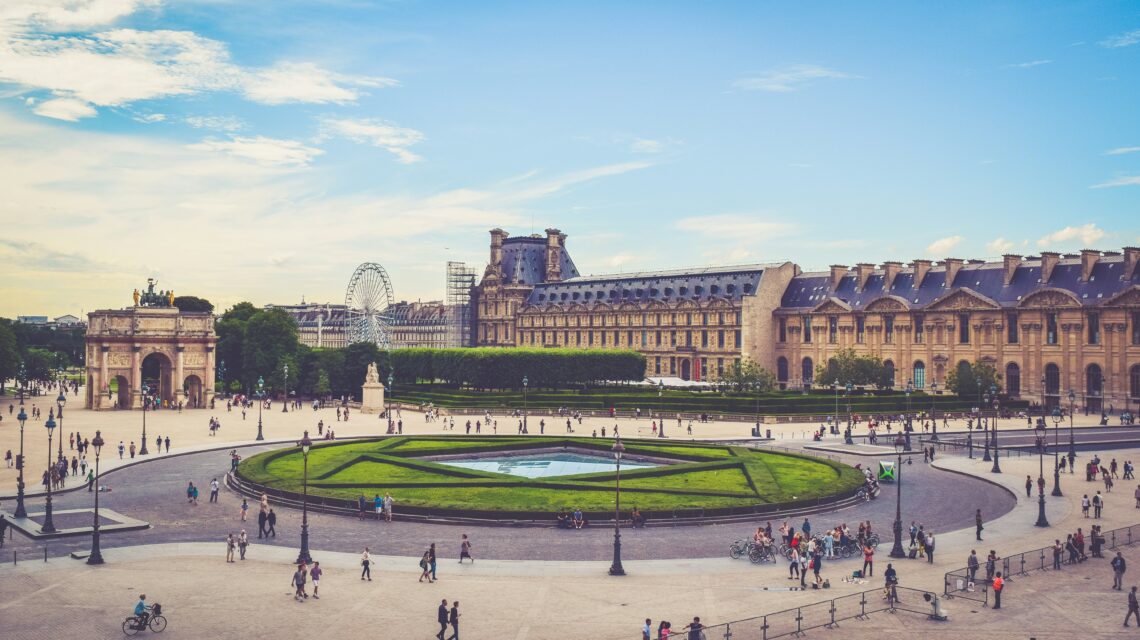 Wide plaza scene with the Louvre Palace in the background, surrounded by greenery. People walk and cycle under a bright blue sky with scattered clouds.