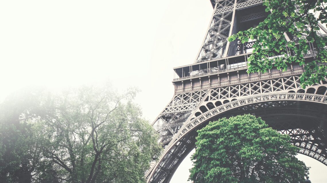View of Eiffel Tower base amidst lush green trees, framed by a hazy, overcast sky. The scene conveys a sense of grandeur and tranquility.