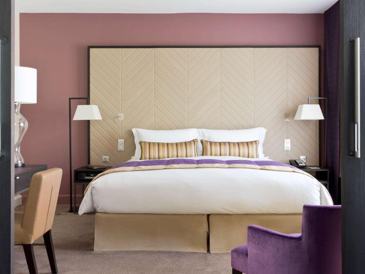 Elegant hotel room with a king-sized bed against a beige herringbone headboard, flanked by modern lamps. Purple accents and a stylish armchair add warmth.