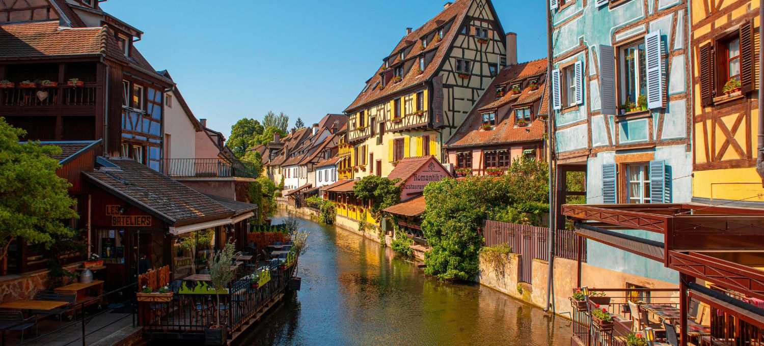Charming canal scene in a colorful European village, featuring half-timbered houses, vibrant facades, and a serene waterway. The sunny day adds warmth.
