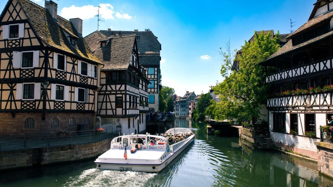 A scenic boat glides along a canal in a quaint European town, surrounded by charming timber-framed houses. The setting is peaceful and idyllic under a bright blue sky.