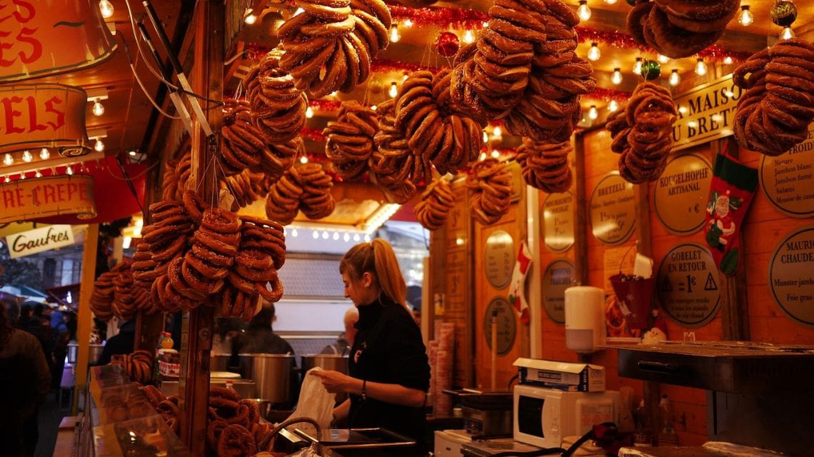 A bustling market stall with golden lights displays numerous hanging pretzels. A vendor serves snacks, surrounded by festive, warm-toned decor.