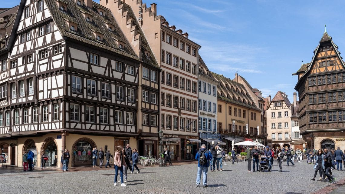 A bustling European square with people walking on cobblestones, surrounded by half-timbered buildings and shops under a clear blue sky, conveying vibrancy.