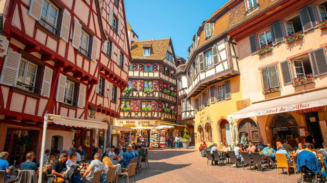 Charming European square with colorful half-timbered buildings. People dine at outdoor cafes, enjoying a sunny day. Lively and picturesque atmosphere.