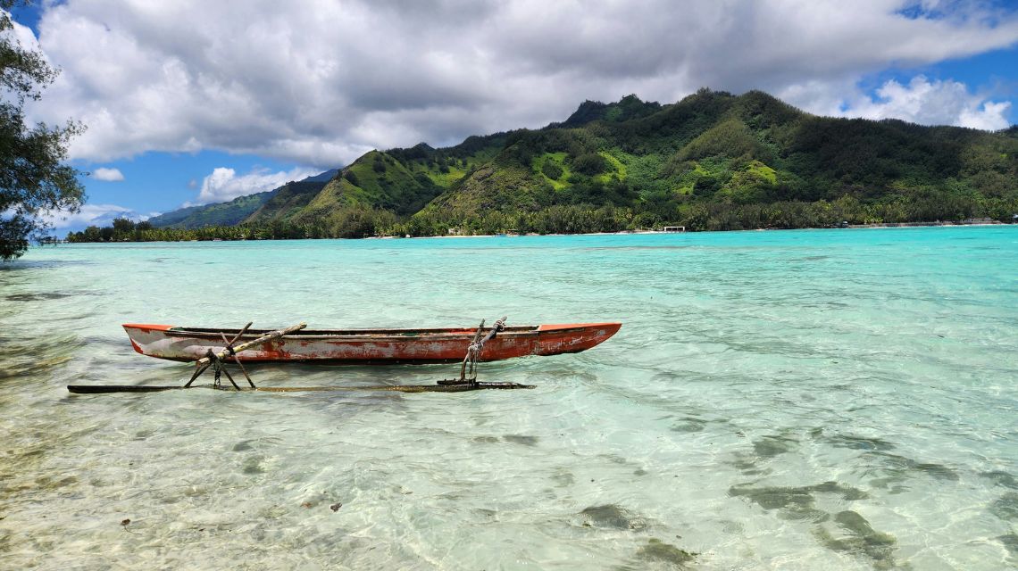 A weathered canoe rests on clear, shallow turquoise water with a backdrop of lush green mountains under a partly cloudy sky, evoking tranquility.
