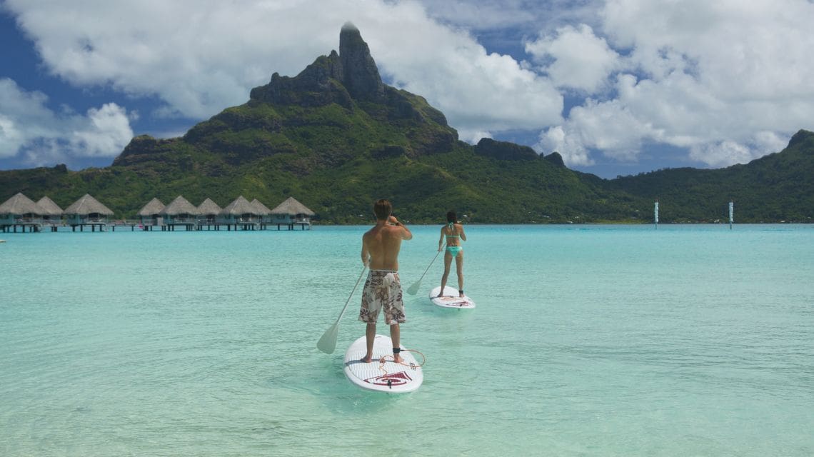 Two people paddleboarding on clear turquoise water, set against a backdrop of lush green mountains and overwater bungalows under a partly cloudy sky.