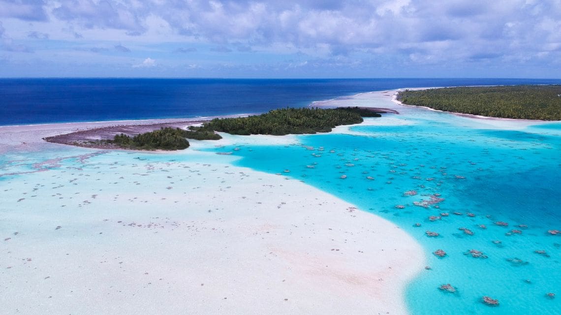 A stunning aerial view of a tropical island surrounded by vibrant turquoise water and white sandy beaches, under a partly cloudy sky. Lush greenery lines the horizon.