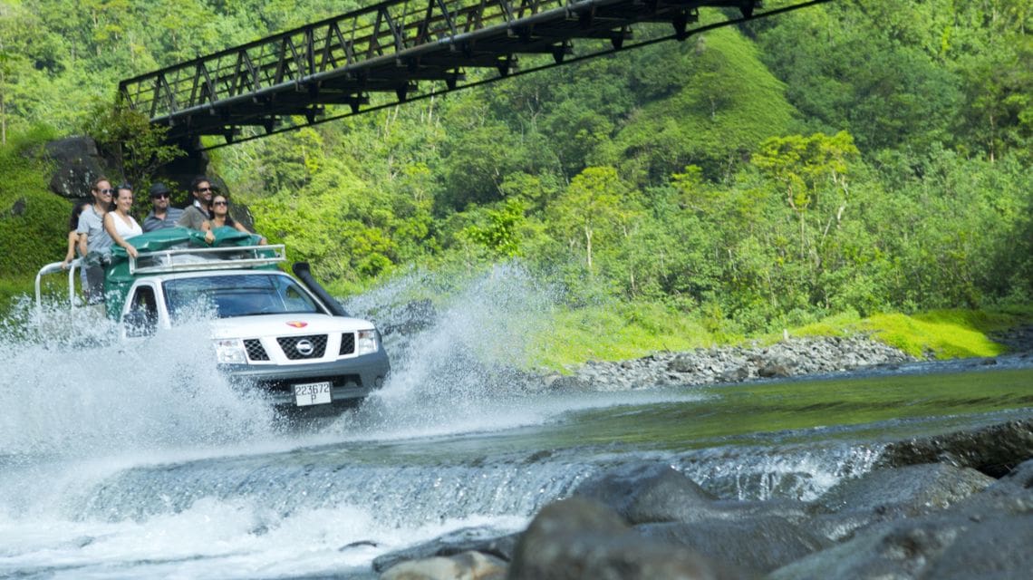 A white SUV crosses a shallow river, splashing water, with people on top enjoying a scenic ride. Lush green forest and a metal bridge are in the background.