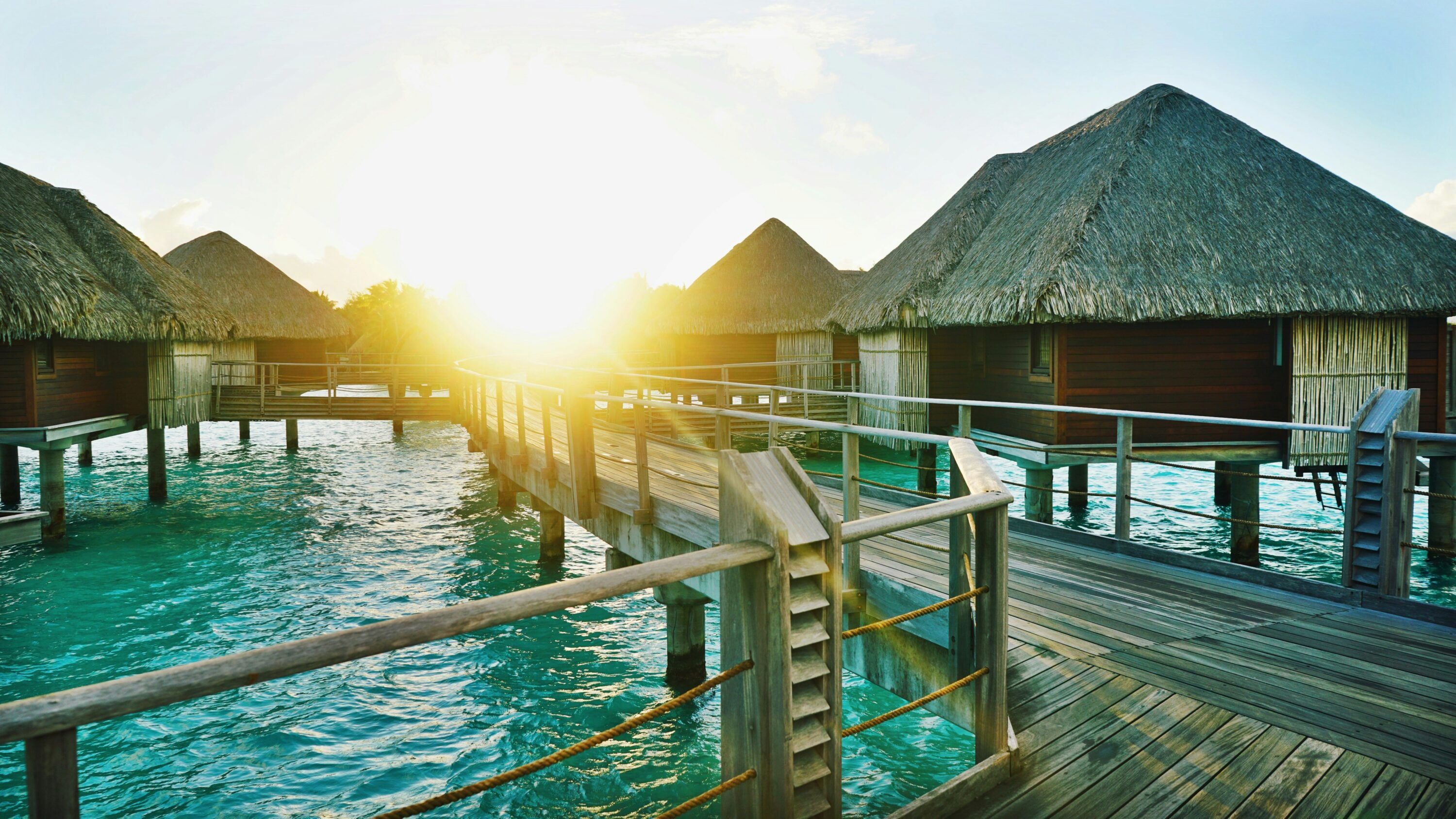 Sunlit overwater bungalows with thatched roofs on turquoise water, connected by a wooden walkway. A serene and tropical island atmosphere at sunset.