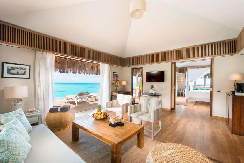 Luxurious beachfront villa interior with wooden floors, cozy seating, and tropical decor. Open doors reveal a deck with lounge chairs facing the ocean.