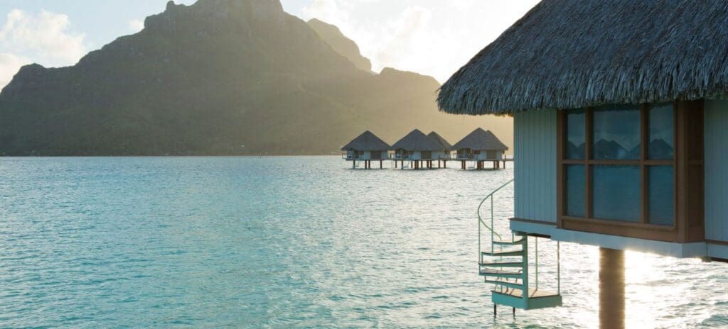 Overwater bungalows with thatched roofs sit on turquoise water, a mountain in the background. The serene setting conveys tranquility and tropical beauty.