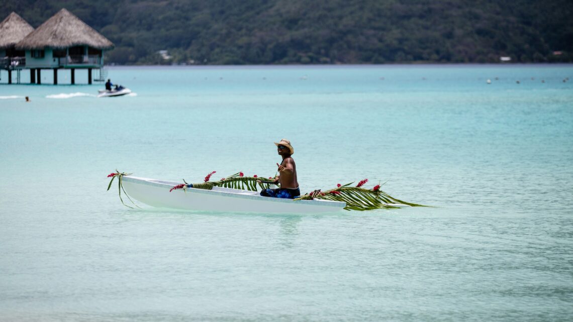 A person sits in a small, floral-adorned canoe on clear turquoise waters, wearing a straw hat. Overwater bungalows and lush hills are visible in the background.
