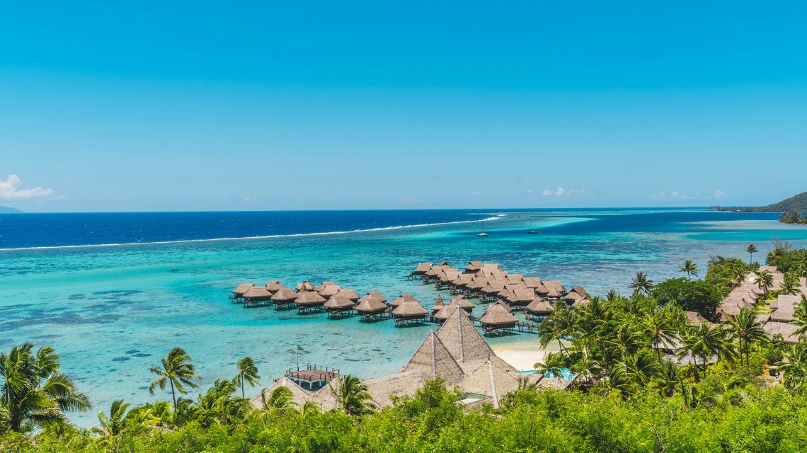 Scenic view of turquoise ocean with overwater bungalows, surrounded by lush palm trees under a clear blue sky, conveying a tropical paradise vibe.