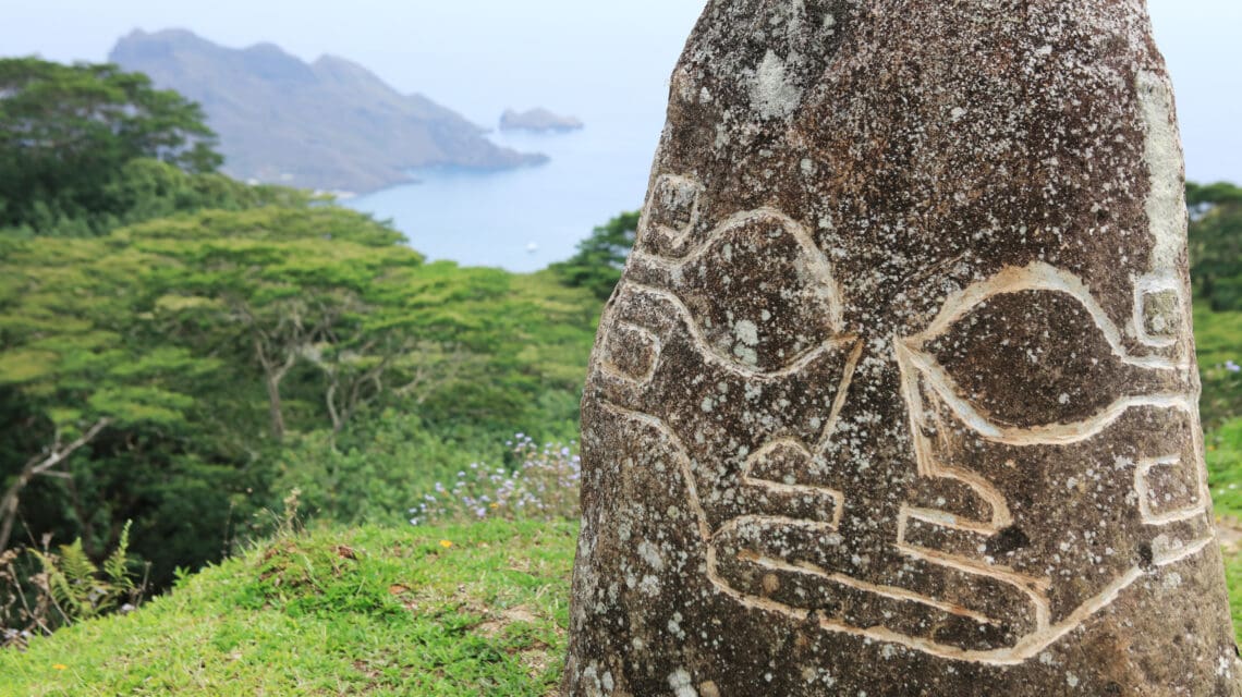 Stone carving with abstract face designs set against lush green foliage and distant ocean hills. The scene is serene and mysterious.