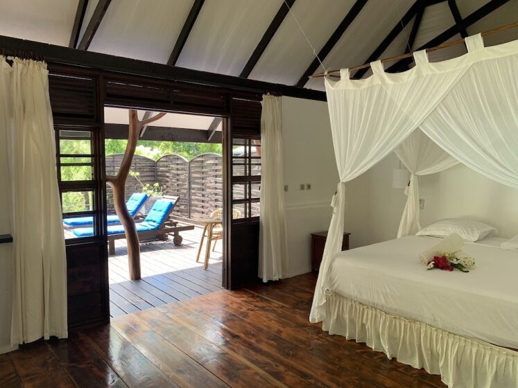 Bedroom with white canopy bed and fresh flowers. Open sliding doors reveal a patio with two blue lounge chairs, surrounded by greenery. Airy and serene.