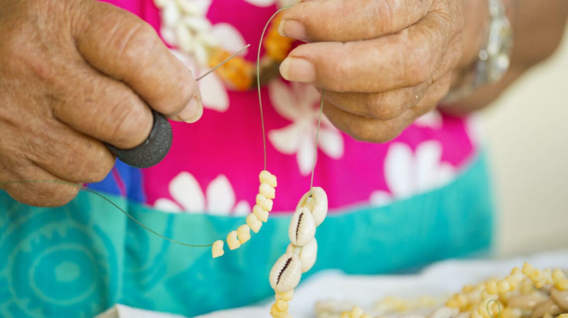 Hands stringing small beads and cowrie shells on a wire. The background shows a pink and turquoise floral garment. The scene is calm and detailed.