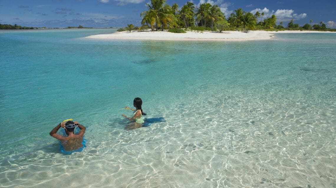 Two people enjoy snorkeling in clear, shallow turquoise water near a tropical island with white sand and palm trees. The scene is serene and sunny.