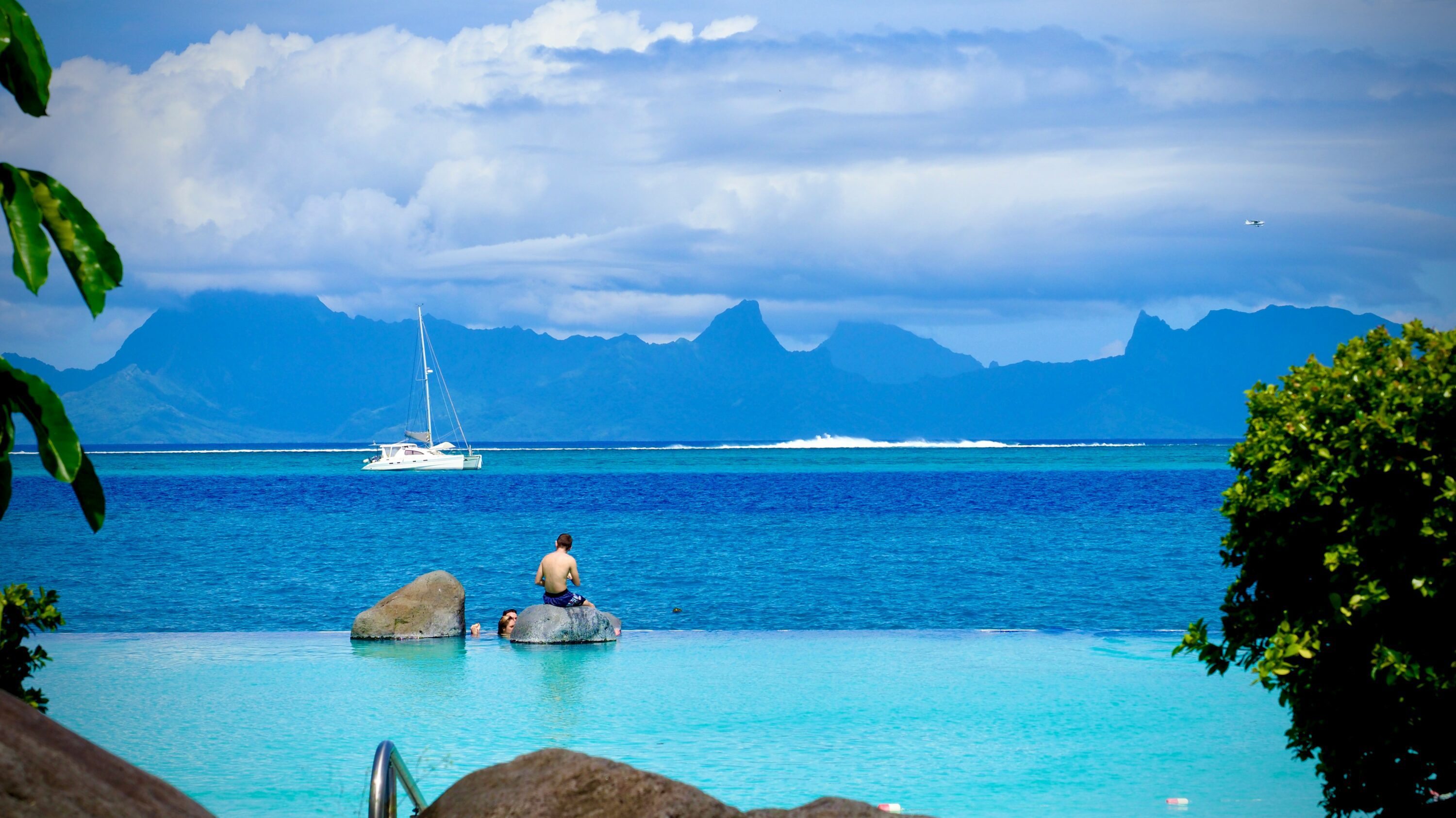 A person sits on a rock in a serene blue pool, overlooking the ocean with a distant sailboat and mountain range under dramatic clouds. Peaceful, tropical vibe.