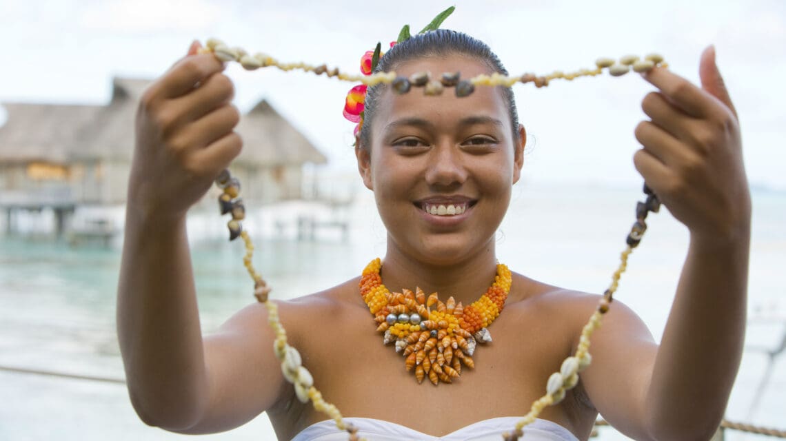 Smiling woman wearing a shell necklace, holding another shell lei, with tropical huts over water in the background. Vibrant, joyful atmosphere.