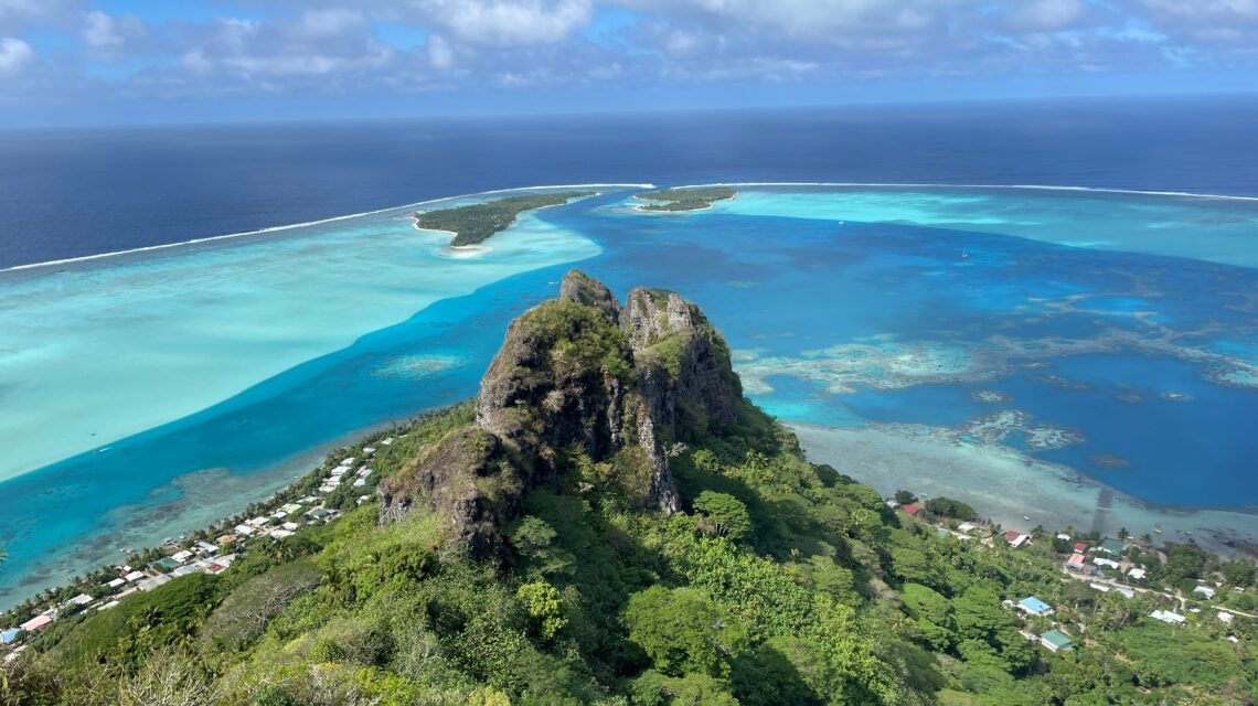 Aerial view of a lush green volcanic landscape in Maupiti, French Polynesia. Vibrant turquoise lagoon contrasts with deep blue ocean under a partly cloudy sky.