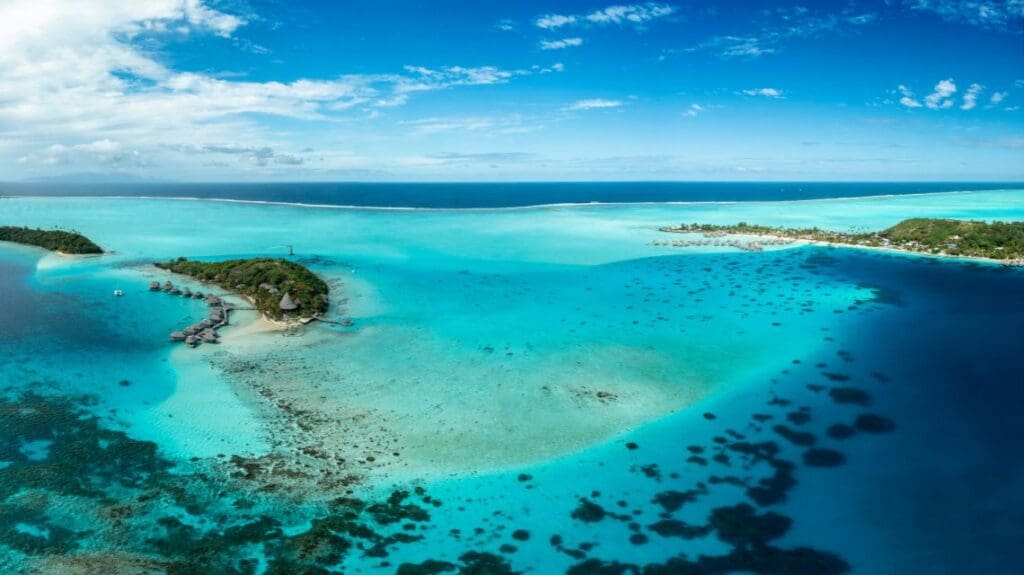 Aerial view of a tropical island with lush greenery surrounded by vibrant turquoise water. Overwater bungalows extend into the sea, under a clear blue sky.