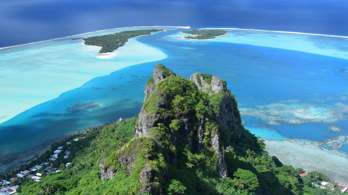 Aerial view of a tropical island with lush green mountains in the foreground. Surrounding turquoise and deep blue ocean waters create a serene, vibrant scene.