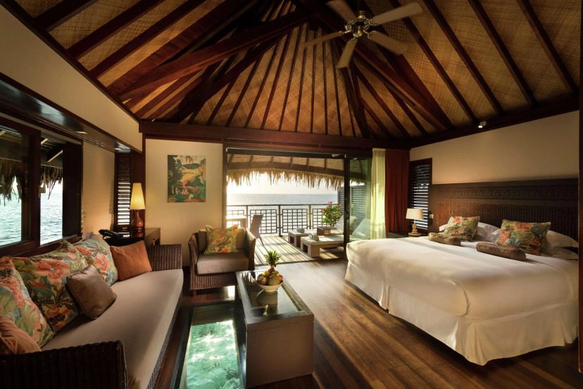 Luxurious tropical villa with a thatched ceiling, wooden floors, plush sofa, large bed, and balcony overlooking the water, creating a serene ambiance.