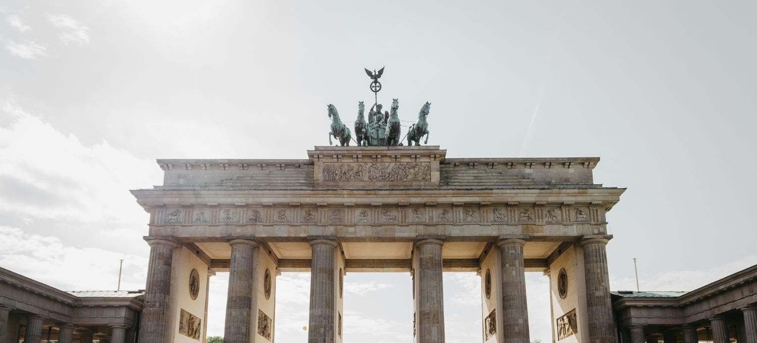 The image shows the Brandenburg Gate in Berlin, a neoclassical monument with tall columns and a chariot sculpture atop, set against a cloudy sky.