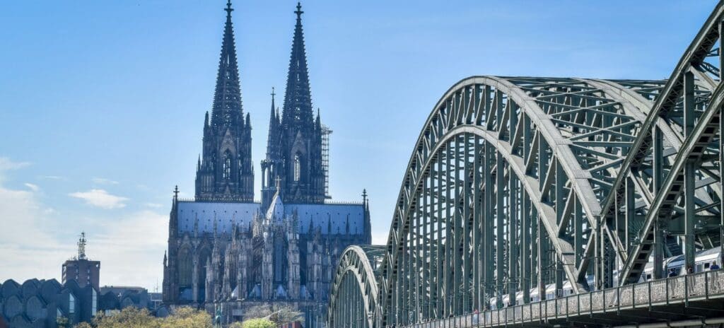 Cologne Cathedral's twin spires rise majestically against a clear blue sky, with the Hohenzollern Bridge's arched structure in the foreground.