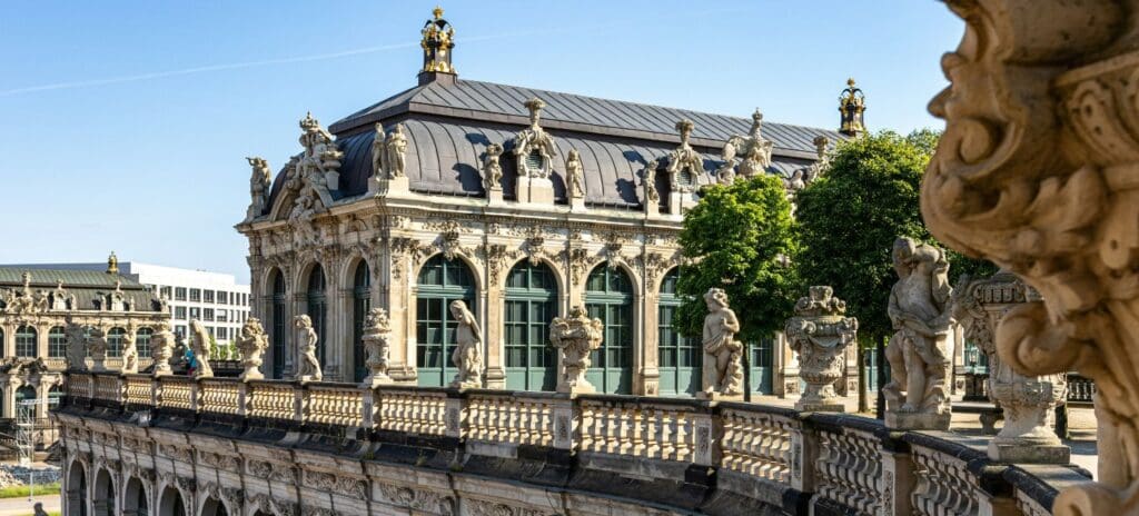 Ornate Baroque palace with arched windows and statues on a balustrade, set against a clear blue sky with lush green trees, conveying elegance and history.