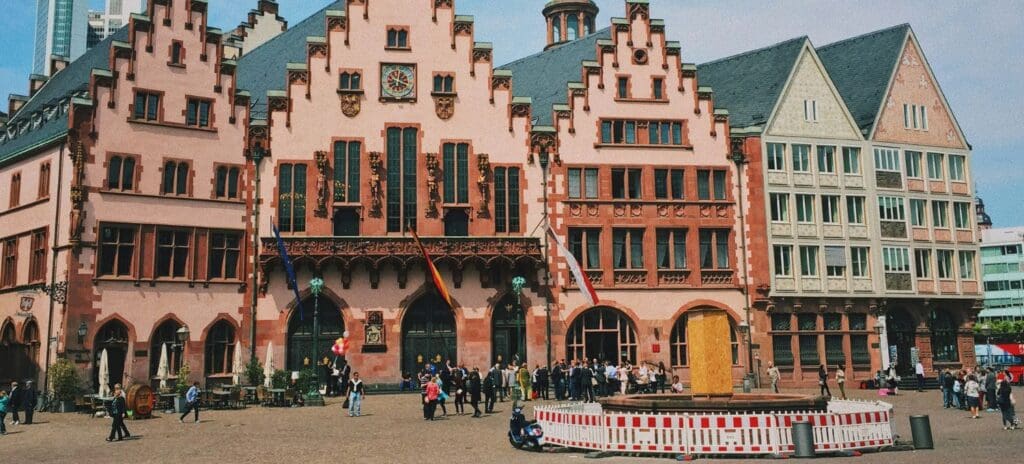 Historic Römer building in Frankfurt with stepped gables and flags. A square in front hosts people and a barricaded area with a fountain. Vibrant, lively scene.