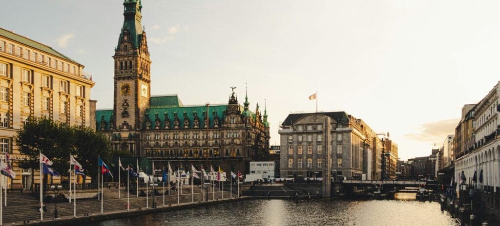 Historic building with a green roof overlooking a canal at sunset. Flags line the water, and surrounding structures reflect golden light, creating a peaceful, nostalgic atmosphere.