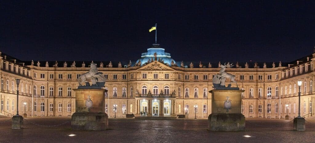 A grand, illuminated palace at night, with intricate architecture and a central dome with a flag. Statues on pillars flank the entrance, creating a regal ambiance.