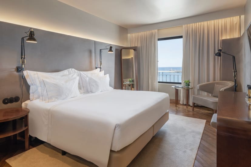 Elegant hotel room with a large bed, soft lighting, and a window view of the sea. Cozy atmosphere with neutral tones, a chair, and a full-length mirror.