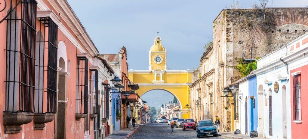 Street view in Antigua, Guatemala, featuring colorful colonial buildings lining a cobblestone road. The iconic yellow Santa Catalina Arch stands prominently.