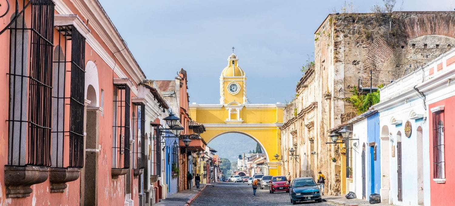 Street view in Antigua, Guatemala, featuring colorful colonial buildings lining a cobblestone road. The iconic yellow Santa Catalina Arch stands prominently.
