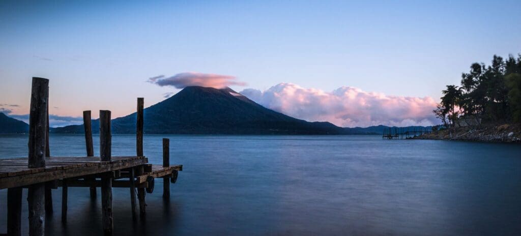 Peaceful scene of a serene lake at dusk with a wooden dock on the left, tranquil water, and a silhouette of a volcano under a pink and blue sky.