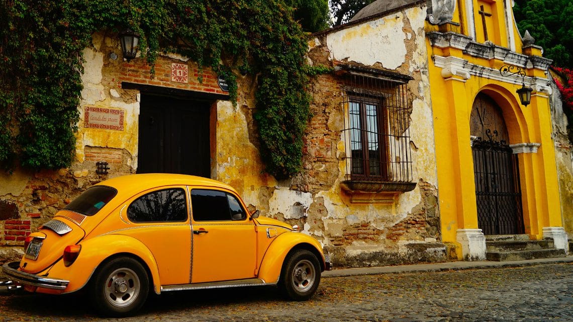 A vintage yellow Beetle is parked on a cobblestone street beside a rustic, weathered building with ivy-covered walls and a bright yellow arched entrance.