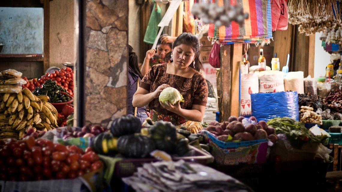 A woman stands in a vibrant market, examining a cabbage. Surrounding her are stacks of bananas, tomatoes, and diverse vegetables, creating a lively, busy atmosphere.