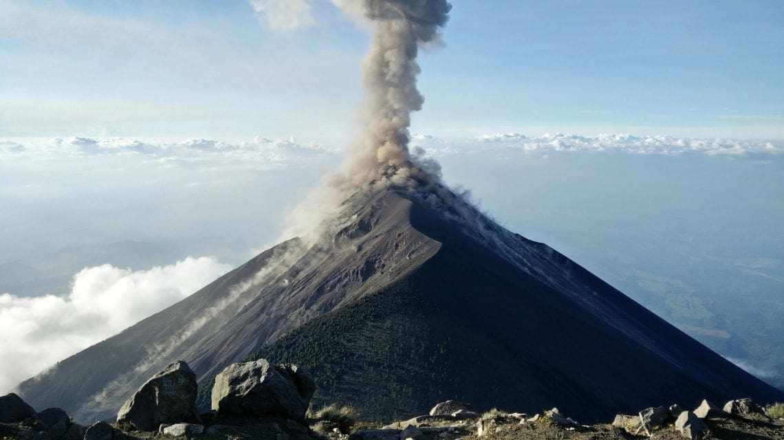 A towering volcano erupts, spewing a thick plume of ash into the clear blue sky. Surrounding rocks and distant clouds add dramatic contrast.