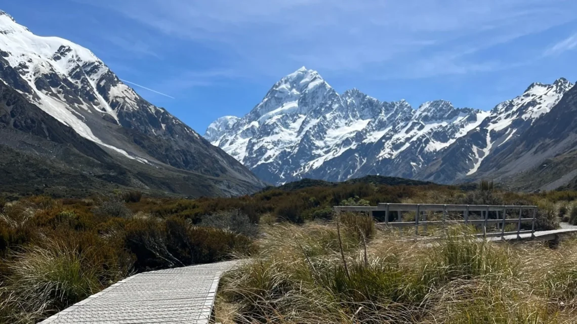 Boardwalk through rugged scrub brush with jagged snow-capped mountains in the background.