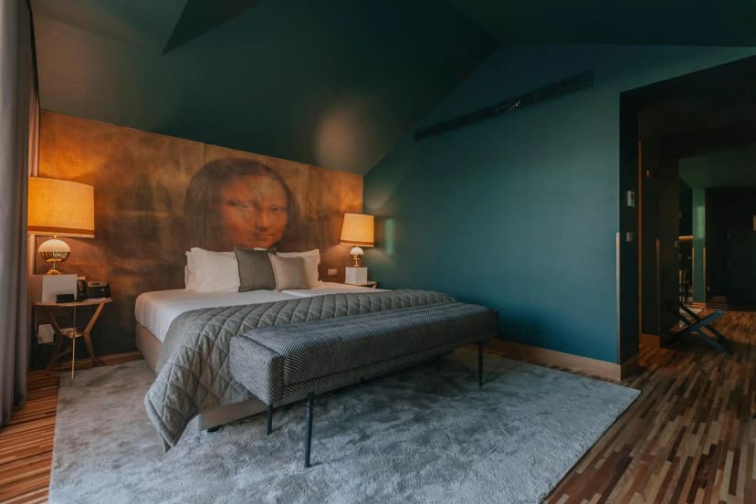 A cozy bedroom with teal walls features a large print of the Mona Lisa above the bed. Warm lighting from table lamps creates a calm atmosphere.