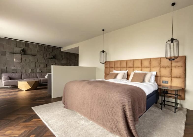 Modern bedroom with a cozy double bed featuring a brown quilt, padded headboard, and pendant lights. A stone accent wall and gray sofa add elegance.
