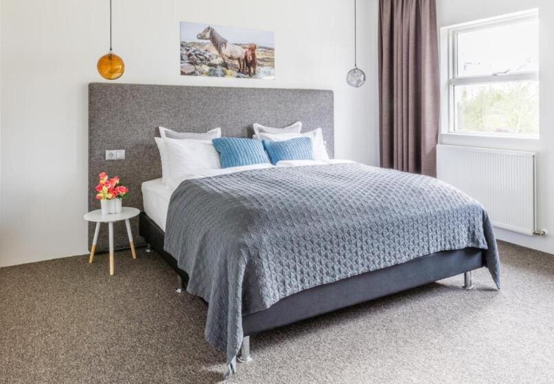 Modern bedroom with cozy queen-sized bed featuring gray quilt and blue pillows. Wall art of horses above bed, soft lighting, and window view.