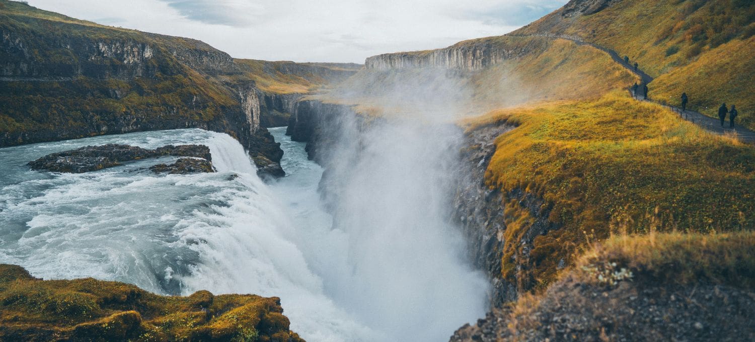 Rushing waterfall cascades into a deep canyon, surrounded by vibrant green and yellow moss-covered cliffs. Mist rises, with small figures on a distant path.