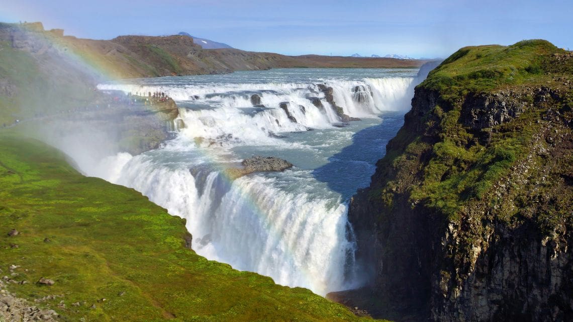 A powerful waterfall cascades down rocky cliffs with lush green grass. A colorful rainbow arches over the mist, creating a vibrant and majestic scene.